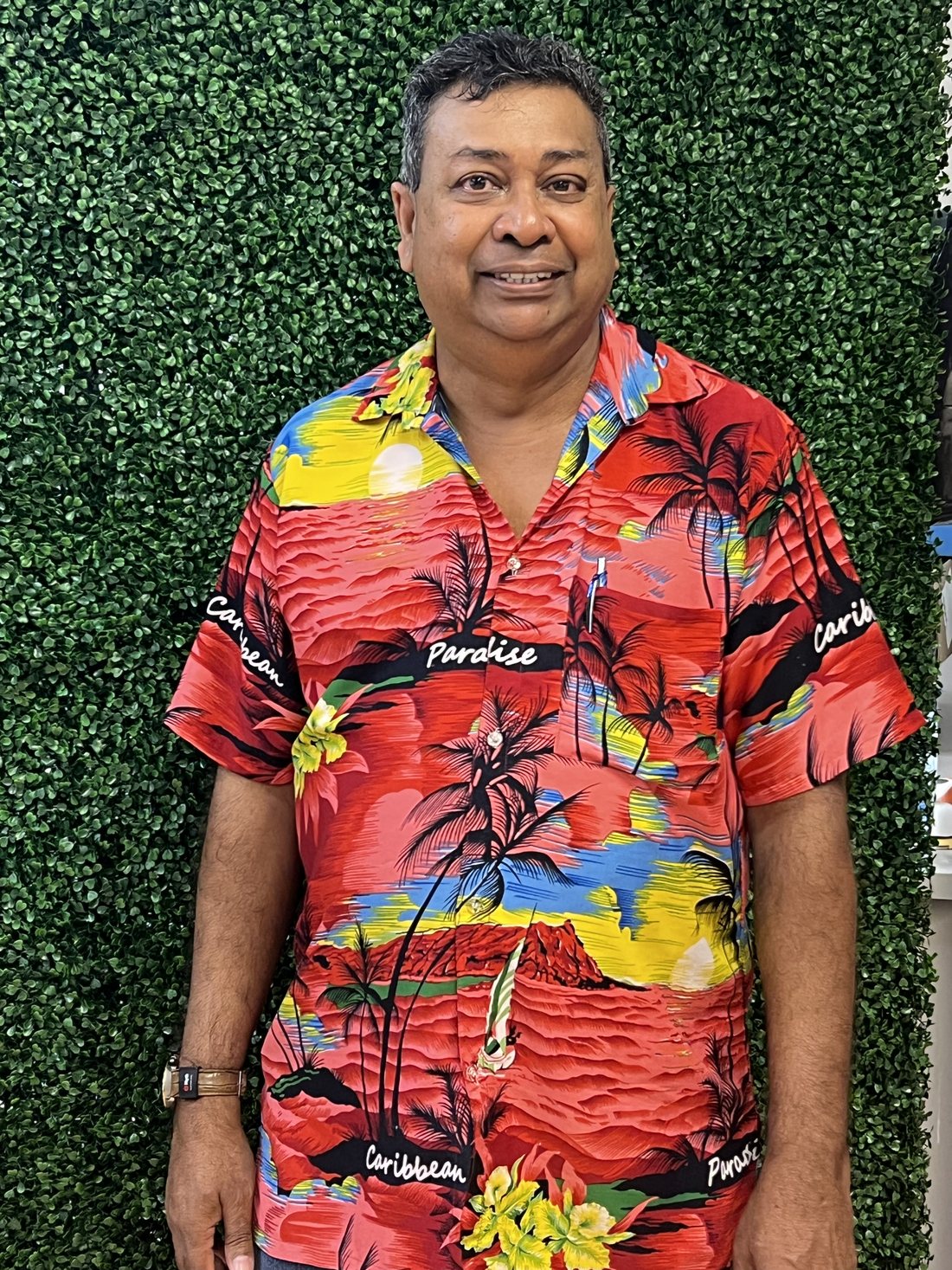 Club member Jameer Ali, in a bright patterned shirt, stands smiling in front of a green foliage wall