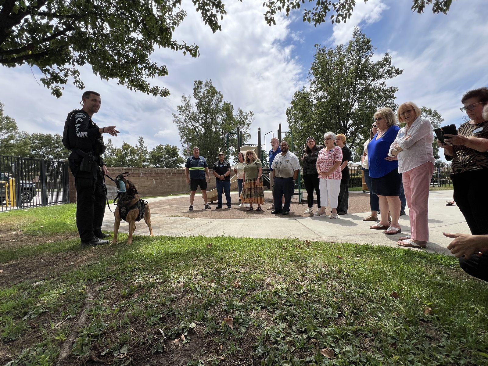 A Beaumont Police corporal holds a K9 on a lead and speaks to about fifteen Rotarians standing on a walkway near a playground