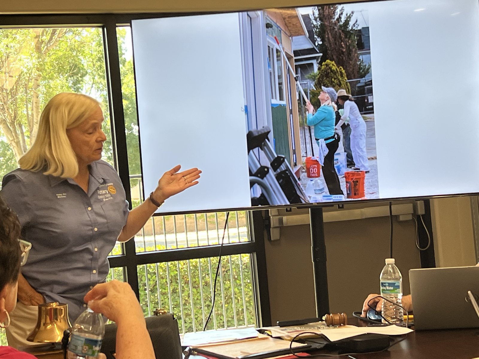 Patsy Orr, in a Rotary District 5330 shirt, gestures toward a screen showing volunteers painting a house during a Rotary service project